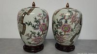Pair of Chinese ceramic urns side by side showing detailed floral hand painted artwork and wooden bases.
