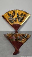 Two Chinese floral design fans, one folded on a surface and the other fully open, showing the gold tonal surface with painted flowers in multiple colors and reddish brown wooden ribs.
