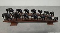 Two sets of hand carved ebony elephant figurines arranged in a line on wooden bridge bases, displayed on a gray floor against a neutral wall.