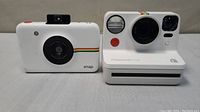 Two instant cameras side by side showing front views with lens, flash and shutter button visible