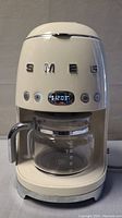 Front view of coffee maker showing cream plastic body, chrome SMEG logo, LED panel lit and glass carafe in place