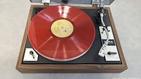 Photo showing full turntable with red record applied to platter, wood finish base, and controls on the right side.