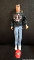 Front view of the Danny doll standing upright wearing black sweatshirt, acid-wash jeans, white shoes, with red baseball cap placed on the ground in front.