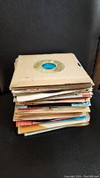 Stack of about 50 vintage 45rpm vinyl records in worn paper sleeves, stacked vertically.