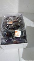Box of multiple pairs of new sunglasses individually packaged in plastic with orange 'balli' tags visible on most.