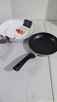 Photo showing both frying pans, one black with wear on handle and one white new pan with packaging
