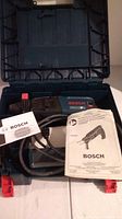 Open case showing Bosch Bulldog rotary hammer drill, power cord, and two manuals inside case compartments.