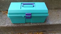 Closed light blue plastic toolbox with purple handle and latch shown from the front on concrete steps
