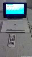 Toshiba portable DVD player powered on with screen showing 'Welcome'. Device open with DVD tray visible, power cord plugged in, remote control placed in front.