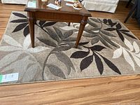 Brown toned rectangular area rug with leaf motifs in beige, cream, taupe, and dark brown tones laid on a wooden floor beneath a wooden table.