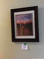 Framed print of a lighthouse on rocky cliffs at sunset, signed by Valerie Chessell, with a dark wood frame and white matting.