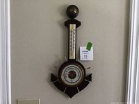 Full view of the wall-mounted barometer and thermometer item against a beige wall, showing entire wooden body shaped like ship's wheel and anchor.