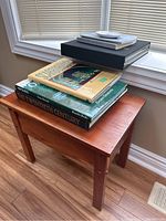 Small wooden bench with a stack of assorted coffee table books on top, placed near a window on laminate floor.