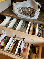 Open drawer showing mixed metal spoons, forks, and knives alongside various kitchen utensils such as peelers and tongs, plus a plastic bag filled with extra spoons and forks on countertop.