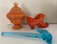 Three glass items on shelf: orange slag glass covered candy dish, orange slag glass shoe figurine, and blue Vaseline glass tomahawk decanter.