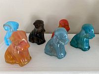 Six colorful Degenhart glass dog figurines arranged in two rows on a shelf, showing variety of colors including blue, orange, red, black, and teal.