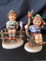 Front view of two Hummel figurines: Wayside Harmony boy sitting on wooden fence with bird, and Apple Tree Boy sitting on apple tree branch.