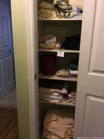 A closet with four shelves containing folded sheets, towels, blankets, and a red bathroom waste basket.