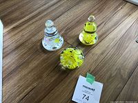 Three glass paperweights on a wooden surface: two bell-shaped with clear glass and yellow floral designs, one round with yellow floral design in clear glass.