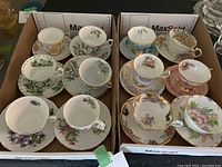 Box with 12 assorted vintage fine china teacups and saucers separated into compartments showing various floral designs and gold trim.