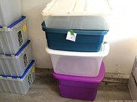 Stacked four plastic storage totes with lids in colors clear, teal blue, translucent white, and purple. The totes are in four different sizes, stacked largest to smallest from bottom to top.
