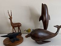 Six carvings displayed together: a large wooden dolphin and whale, a slender gazelle, a yellow painted bird, an unpainted wood bird, and a small black cast iron anvil on wood base.