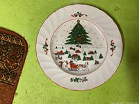 Top view of one Christmas-themed dinner plate showing decorated Christmas tree, village scene, horse sleigh, and holly accents with red rim