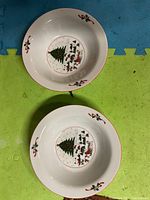 Top view of two white ceramic serving bowls showing Christmas tree and gift design at the center with holly and red ribbons on the rim.