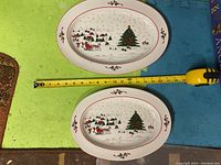 Top view of two oval Christmas-themed serving platters with holiday tree and village design, taped measure shows larger platter about 15 inches.