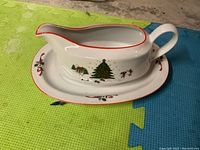 Christmas-themed porcelain gravy boat with handle and attached underplate, white with red rim and decorated with a Christmas tree and festive characters