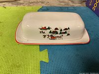 Side view showing the Christmas-themed holiday design on the lid with horses, sleigh, village, and trees, and red border around the dish.