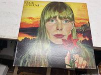 Front cover of Joni Mitchell Clouds album showing painted portrait of the artist holding a red flower with sunset background.