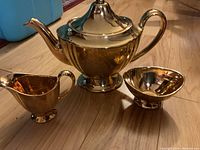 Gold-tone Royal Winston teapot, creamer, and sugar bowl arranged on wooden floor.