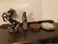 Two clocks and two figurines on a wooden surface against a white background. One figurine is a rearing horse with detailed mane and tail. The other is a figurine of a kneeling person. Both clocks are wooden framed, one octagonal.