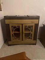 Front view of painted wood cabinet showing two glass doors with decorative metalwork and two drawers, in brown and gold finish.