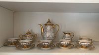 Full espresso set showing teapot, creamer, sugar bowl, six matching cups and saucers, all white porcelain with intricate gold floral detailing and gold rims.
