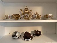 Full view showing Japanese porcelain tea set with floral and gold designs on the top shelf and six brown ceramic espresso cups and saucers by ACF, on lower shelf