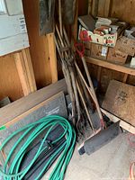 Multiple wooden-handled shovels, rakes, and a push broom stacked inside a wooden shed.