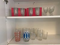Three sets of glasses on a white shelf inside a cabinet. One set of 4 clear glasses on a red cardboard backing, two vintage style goblets, and assorted other glasses including a blue oval decorated glass and an orange striped glass.