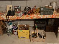 Wide view showing the hardware storage organizer, extension cords, multiple phone handsets, paint cans and various small tools on a concrete shelf with floral tablecloth underneath.