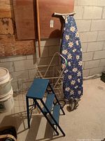 Image of the folding stepladder, ironing board with floral cover, and folding metal shopping cart.