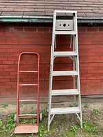 Front view of aluminum folding ladder and red metal dolly leaning against a brick wall