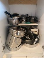 View inside cabinet showing stacked stainless steel pots, pans, and pressure cooker with black handles and lids.