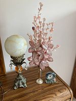 Vintage lamp with ornate metal base, coloured glass globe, metal vase with pink faux flowers, and decorative clock with dolphins on wood surface.