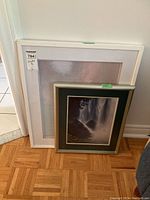 Two framed prints leaning against a door: smaller waterfall print with dark green mat and white frame in front, larger pale abstract print with white frame behind