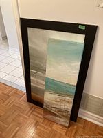 Two wall prints leaning against a wall on wood floor, one large wooden framed print with gray and silver tones and one narrow blue and brown textured canvas print.