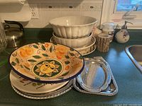 Photo showing assorted serving dishes on a kitchen counter, featuring the large colorful ceramic bowl, the white ceramic bowls stacked, and metal serving trays beneath