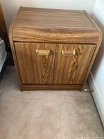 Front view of wooden side table showing two doors and brass handles, on carpeted floor next to wall.