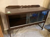 Sideboard shown with glass sliding doors at front, dark natural wood finish with decorative scalloped backsplash, supported by four legs. Visible surface wear and scratches on wood.