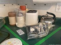 Full lot on kitchen counter includes toaster, kettle, hand mixer beaters, plastic thermoses wrapped in bags.